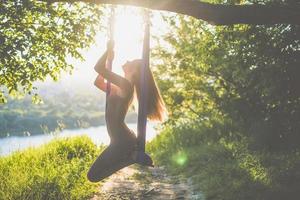 A young female gymnast is engaged in aerial yoga, using a combination of traditional yoga poses, pilates and dance using a hammock at sunset in nature. Healthy lifestyle. photo