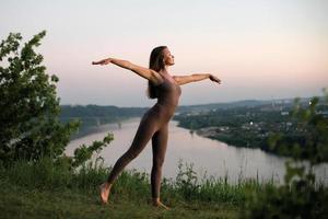 una joven gimnasta hace yoga en la naturaleza en un parque contra el cielo, usando una combinación de posturas de yoga tradicionales, pilates y danza suave. conexión con la naturaleza. foto