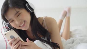 hermosa joven asiática acostada feliz con auriculares escuchando música con relajación y disfrute en el dormitorio, chica divertida y relajada con auriculares tocando canciones mp3 en teléfonos móviles inteligentes, concepto de estilo de vida. video