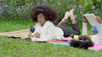 Happy African family with children lying reading or writing to book in the garden, kids go to picnic listening music with headphone, student study and doing homework, education in summer concept. video