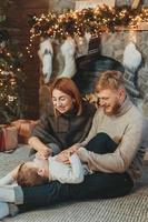 joven familia caucásica mamá papá hijo cerca de la chimenea árbol de navidad foto