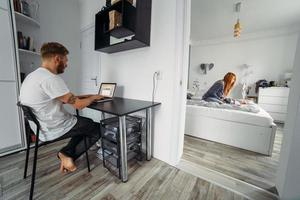 Dad works at laptop, mom and baby on the bed photo