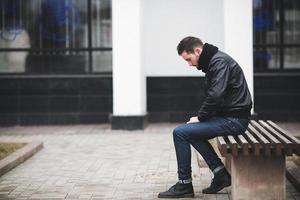 un hombre vestido con jeans y asientos de chaqueta negra en un banco foto