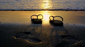 Sandalen am Strand auf einem wunderschönen Hintergrund des Strandes bei Sonnenuntergang mit goldenem Licht, das auf der Wasseroberfläche und weichen Wellen reflektiert wird. sommerferien und reisekonzept. video