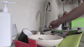 Die Hand eines Mannes öffnet einen Wasserhahn, bevor er in der Küche Geschirr spült. schmutzige Teller, Schüsseln und Tassen in einer Metallspüle. chaotische Küchenszene. fließendes Wasser, Nahaufnahme. Aufräumen nach dem Abendessen. video