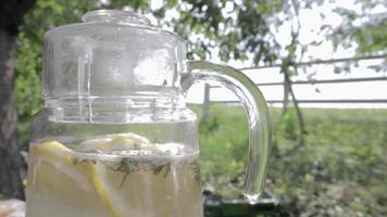 hemgjord lemonad gjord av citroner i en stor glaskanna på bordet i trädgården. en kanna med citron och mynta står på gatan mot bakgrund av grönska en varm sommardag. video