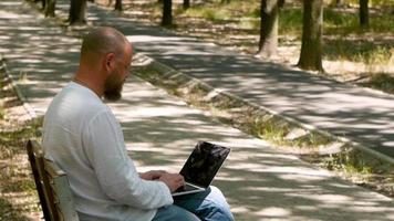 um homem com barba no parque em um banco está digitando texto em um laptop video