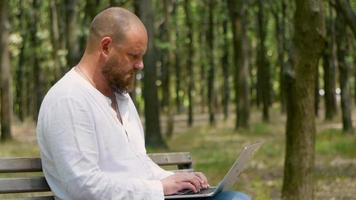 A man with a beard in the park on a bench is typing text on a laptop video