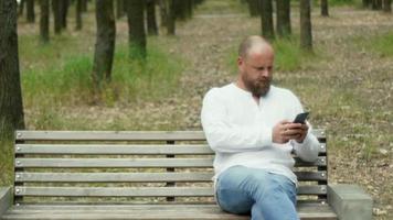 A man with a beard in the park on a bench looks at the phone video