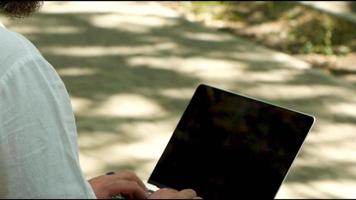 A man with a beard in the park on a bench is typing text on a laptop video