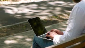 un hombre con barba en el parque en un banco está escribiendo texto en una computadora portátil video