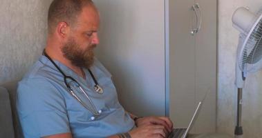 A man with a beard in a doctor's costume on the couch is typing text on a laptop video