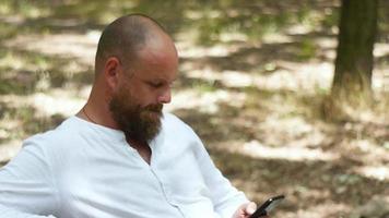 A man with a beard in the park on a bench looks at the phone video