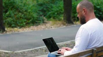 un hombre con barba en el parque en un banco está escribiendo texto en una computadora portátil