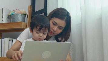 la madre y el hijo asiáticos se sientan en casa y trabajan en su computadora portátil en el trabajo. mamá trabaja en línea hijo se sienta en su regazo. video