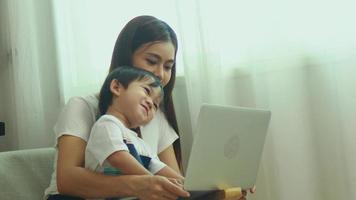 la mère et le fils asiatiques sont assis à la maison et travaillent sur leur ordinateur portable au travail. maman travaille en ligne, son fils est assis sur ses genoux. video