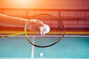 Badminton racket and old white shuttlecock holding in hands of player while serving it over the net ahead, blur badminton court background and selective focus photo