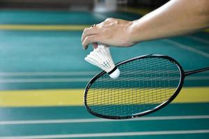 Badminton racket and old white shuttlecock holding in hands of player while serving it over the net ahead, blur badminton court background and selective focus photo