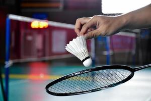 la raqueta de bádminton y el viejo volante blanco sosteniéndose en las manos del jugador mientras lo sirven sobre la red por delante, desdibujan el fondo de la cancha de bádminton y el enfoque selectivo foto