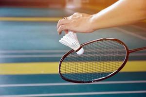 Badminton racket and old white shuttlecock holding in hands of player while serving it over the net ahead, blur badminton court background and selective focus photo