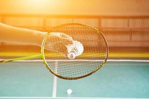 Badminton racket and old white shuttlecock holding in hands of player while serving it over the net ahead, blur badminton court background and selective focus photo