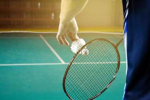 Badminton racket and old white shuttlecock holding in hands of player while serving it over the net ahead, blur badminton court background and selective focus photo