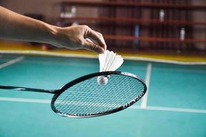 Badminton racket and old white shuttlecock holding in hands of player while serving it over the net ahead, blur badminton court background and selective focus photo