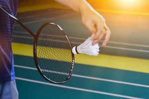 Badminton racket and old white shuttlecock holding in hands of player while serving it over the net ahead, blur badminton court background and selective focus photo