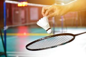 Badminton racket and old white shuttlecock holding in hands of player while serving it over the net ahead, blur badminton court background and selective focus photo