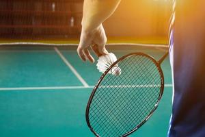 Badminton racket and old white shuttlecock holding in hands of player while serving it over the net ahead, blur badminton court background and selective focus photo