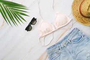 puesta plana de artículos de verano con bikini pastel, gafas de sol, jeans, sombrero y planta tropical verde sobre fondo de mármol, concepto de moda y verano foto