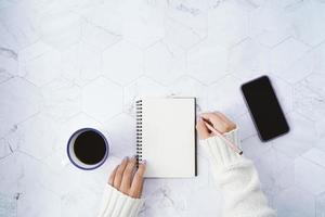 Flat lay of woman writing on blank notebook and drinking hot cup of coffee and using smartphone on white marble background, winter seasonal concept, copy space and mockup photo