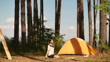 Young woman stretches on a yoga mat outside a yellow tent at camp site