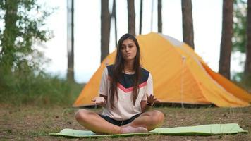 Young woman sits on a yoga mat outside a yellow tent at camp site video