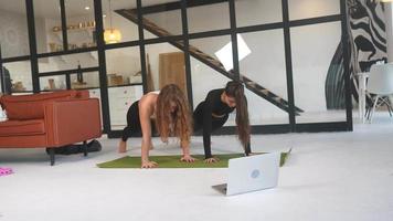 dos mujeres jóvenes hacen ejercicio en una alfombra de yoga en el suelo con una computadora portátil abierta cerca video