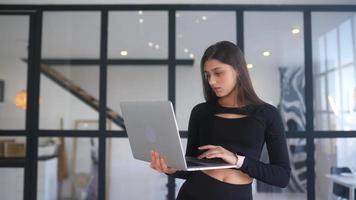 junge Frau in Schwarz steht, während sie einen Laptop in einem offenen Raum mit einem großen Fenster benutzt video
