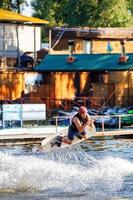 Kyiv. Ukraine. Jun.19.2022. A sportsman-wakeboarder in a protective helmet jumps with a board on the waves over the water surface of the river, holding the rope with his hands. photo