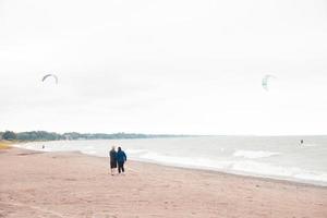 Couple on beach watching kiteboarder on a windy day. Waves crashing into the sandy beach. Whitecaps on the waves. Colorful parasails. Beach sand covered in tracks. Trees lining distant shoreline. photo