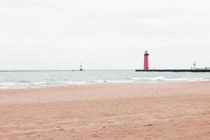 Gray overcast sky of Lake Michigan. Waves rolling into the empty beach. Red lighthouse seen at the end of the pier to warn boaters. Buoys seen in the distance. Footprints in the sand. photo