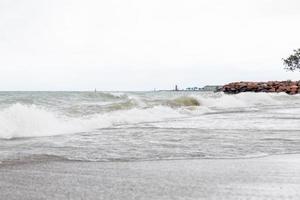 Large waves rolling into the sandy shoreline off Lake Michigan on a cloudy overcast day. Rocks holding the shoreline back with trees in the corner. Buildings and lighthouse seen in the distance. photo