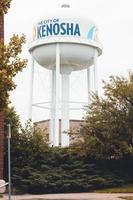 Kenosha, Wisconsin, water tower with  trees around the base. Cloudy sky. Fence hidden by the bushes and trees. Brick building to one side. Blue letters on the tower. Sidewalk in front. photo