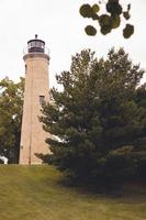 Looking up at an old brick built lighthouse on a cloudy day. Green grass and pine trees surrounding. Windows built in at several levels. Railing around the light on top. photo