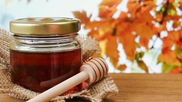 Honey in glass jar with honey dipper on a wooden table with autumn leaves in the background. video