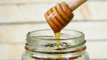 Honey dripping from a honey dipper into a glass honey jar with wood background. video