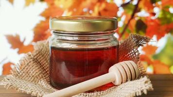 Honey in glass jar with honey dipper on a wooden table with autumn leaves in the background. video