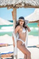 Beautiful brunette girl in a white bathing suit.Portrait of professional swimwear model posing provocatively outdoor.Smiling sensual lady with long legs posing in white bikini and hat the beach. photo