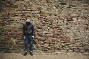 A man dressed in a black jacket and jeans near a stone wall photo