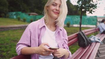 Blonde teen girl sits on a bench while using smart phone and laughing video