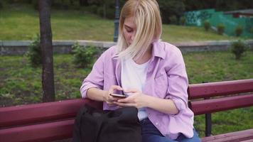 Wind blows the hair of a blonde teen girl sits on a bench while using smart phone and smiling video