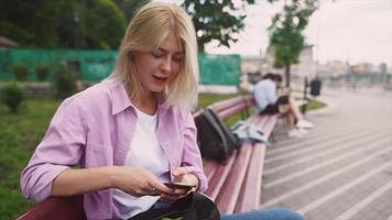 una adolescente rubia se sienta en un banco mientras conversa y usa un teléfono inteligente video